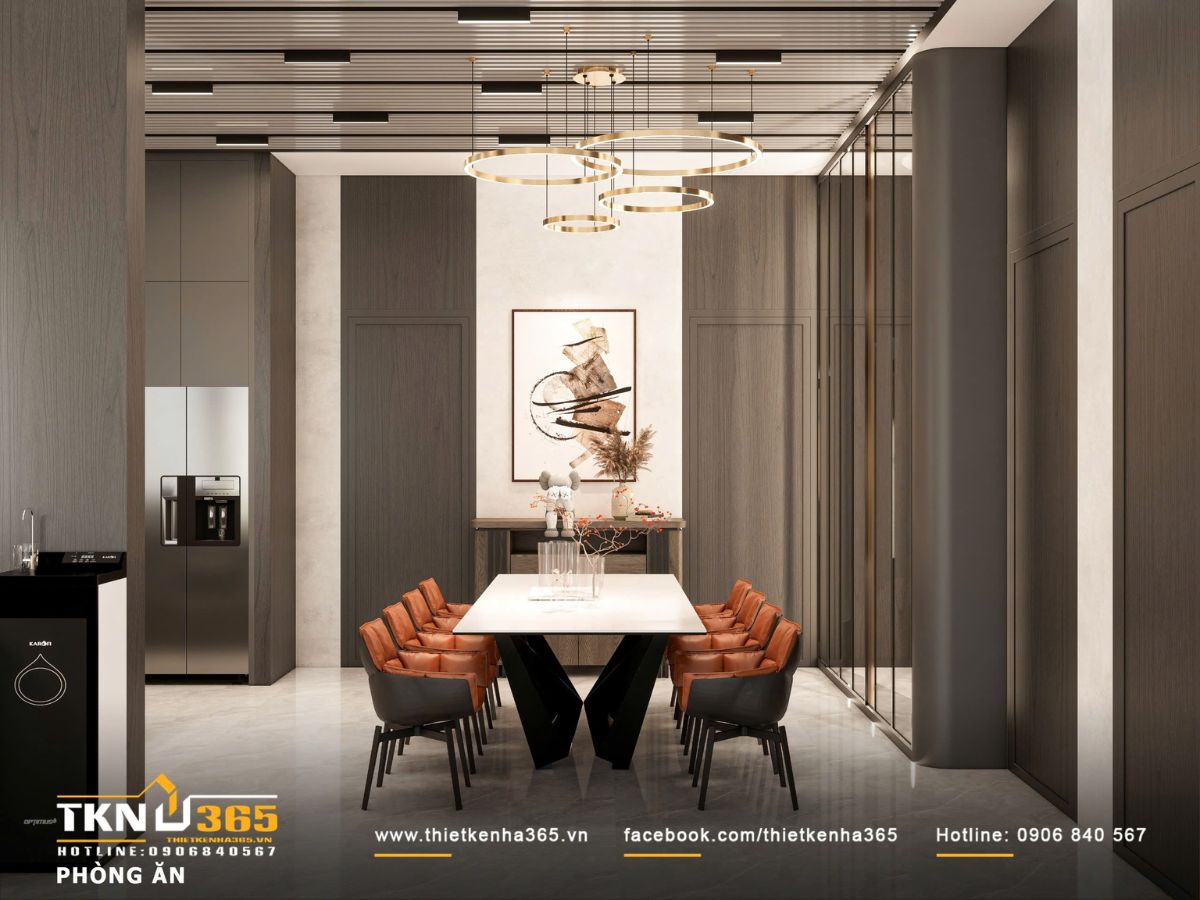 Khu bàn ăn nổi bật với đèn thả vòng ánh kim và nền gỗ tối, tạo điểm nhấn trung tâm ấm áp và sang trọng